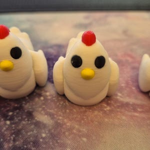 May include: Three white ceramic chicken figurines with red combs and black eyes. The chickens are sitting on a white surface with a galaxy pattern.