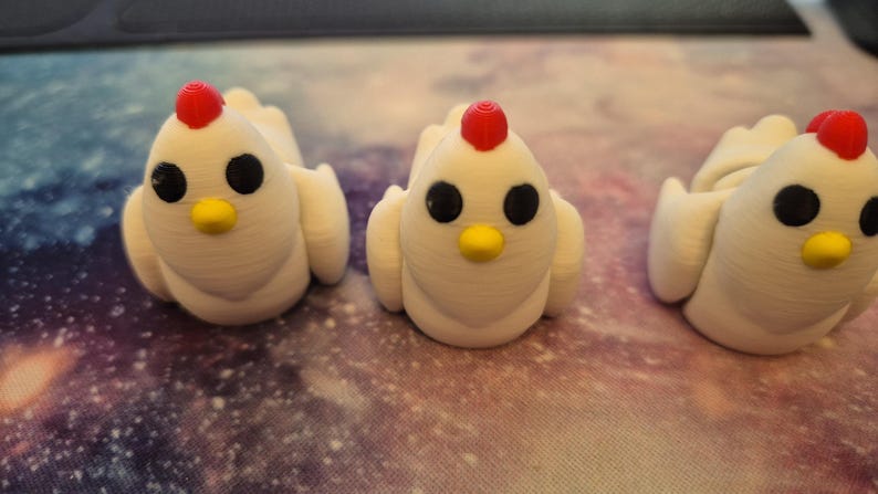 May include: Three white ceramic chicken figurines with red combs and black eyes. The chickens are sitting on a white surface with a galaxy pattern.