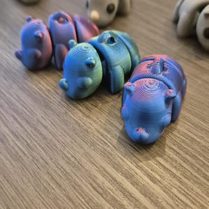 K&ouml;nnte beinhalten: Eine Gruppe kleiner, bunter, 3D-gedruckter Tierfiguren. Die Figuren sind in verschiedenen Blaut&ouml;nen, Gr&uuml;nt&ouml;nen und Violettt&ouml;nen gehalten. Sie sind in einer Reihe auf einer Holzoberfl&auml;che angeordnet.