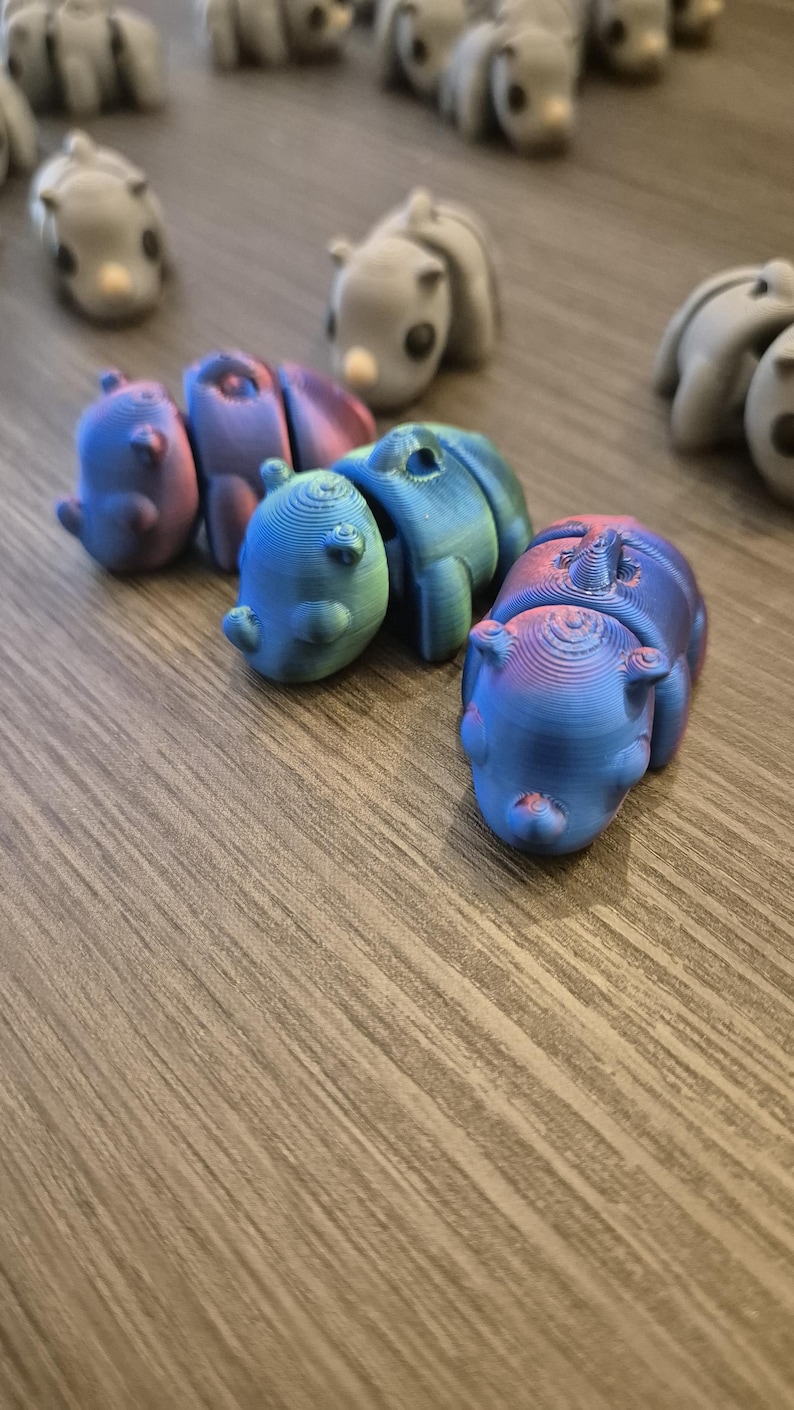 K&ouml;nnte beinhalten: Eine Gruppe kleiner, bunter, 3D-gedruckter Tierfiguren. Die Figuren sind in verschiedenen Blaut&ouml;nen, Gr&uuml;nt&ouml;nen und Violettt&ouml;nen gehalten. Sie sind in einer Reihe auf einer Holzoberfl&auml;che angeordnet.