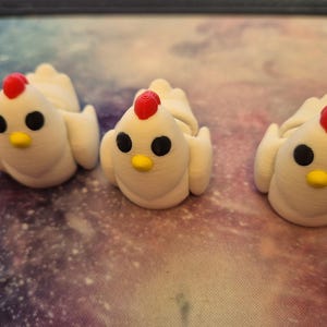 May include: Three white ceramic chicken figurines with red combs and black eyes. The chickens are sitting on a surface with a galaxy pattern.