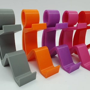 May include: Five 3D printed phone stands in different colors: gray, orange, purple, pink, and red. The stands are shaped like a person sitting with their legs crossed and holding a phone.