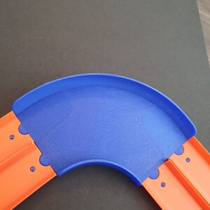 May include: A blue plastic track piece for a toy car racing set. The track piece is curved and connects to two orange track pieces.