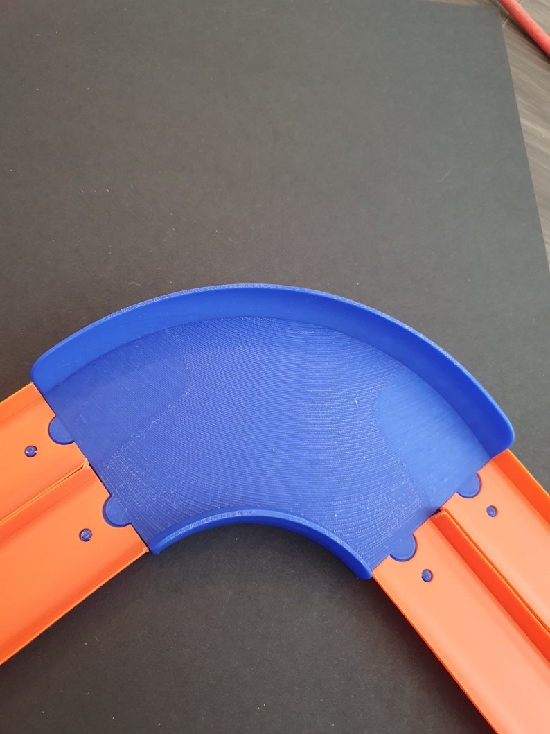 May include: A blue plastic track piece for a toy car racing set. The track piece is curved and connects to two orange track pieces.