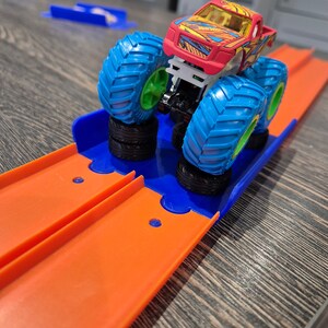 May include: A red, yellow, and blue monster truck toy with large wheels is on an orange and blue plastic track. The truck is on a ramp and has its front wheels in the air.