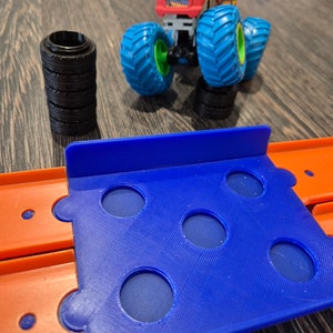 May include: A red and orange monster truck toy with blue wheels and a blue track set with orange sections. The track set has several holes in it.