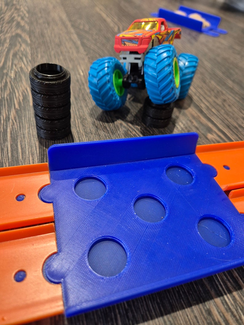 May include: A red and orange monster truck toy with blue wheels and a blue track set with orange sections. The track set has several holes in it.