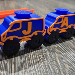 May include: A line of five blue toy trucks with orange accents. Each truck has a letter on the side, spelling out the word "JAXON".