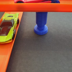 Puede incluir: Dos coches de juguete en una pista de carreras de juguete. La pista superior es naranja y la pista inferior es naranja. Un soporte de pl&aacute;stico azul sostiene la pista superior. El coche de la pista superior es rojo con un parabrisas azul y un dise&ntilde;o de telara&ntilde;a. El coche de la pista inferior es verde y amarillo.