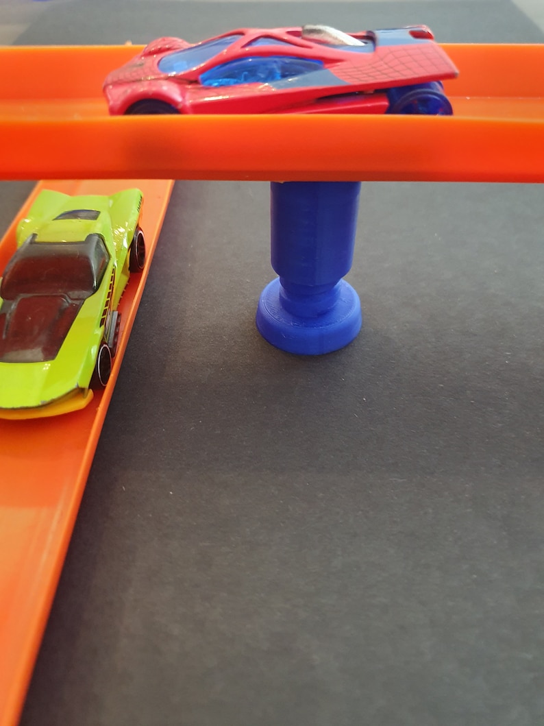 Puede incluir: Dos coches de juguete en una pista de carreras de juguete. La pista superior es naranja y la pista inferior es naranja. Un soporte de pl&aacute;stico azul sostiene la pista superior. El coche de la pista superior es rojo con un parabrisas azul y un dise&ntilde;o de telara&ntilde;a. El coche de la pista inferior es verde y amarillo.