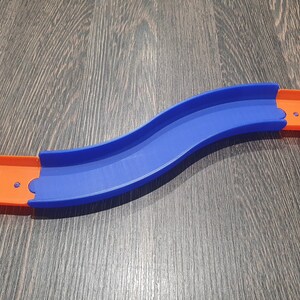 May include: A blue and orange plastic track piece for a toy car race track. The track piece is curved and has a slight bend in the middle.