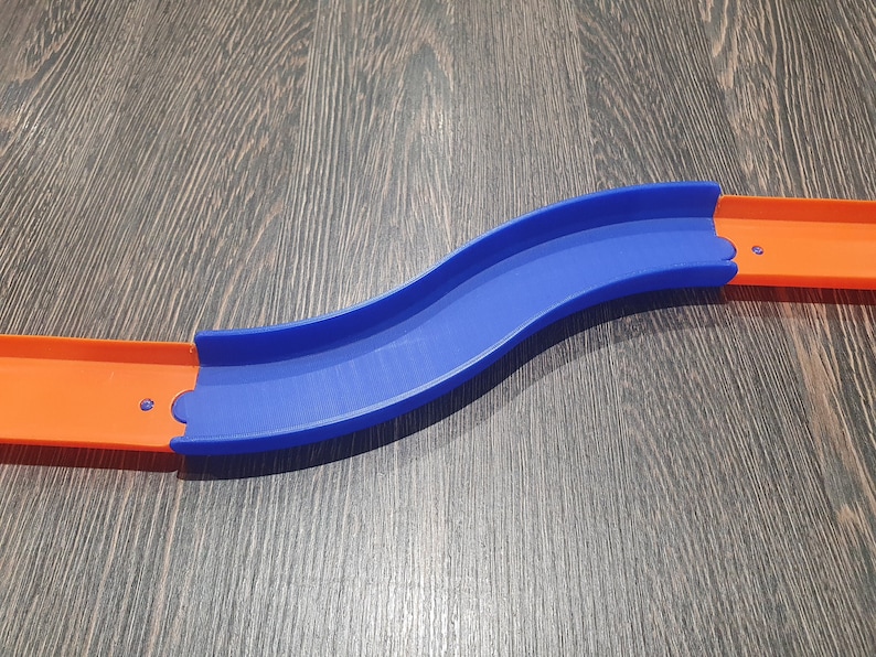 May include: A blue and orange plastic track piece for a toy car race track. The track piece is curved and has a slight bend in the middle.