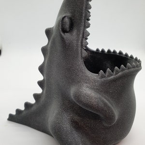 May include: A black, 3D printed dinosaur-shaped planter with a textured surface and an open mouth. The planter is designed to hold small plants or other decorative items.