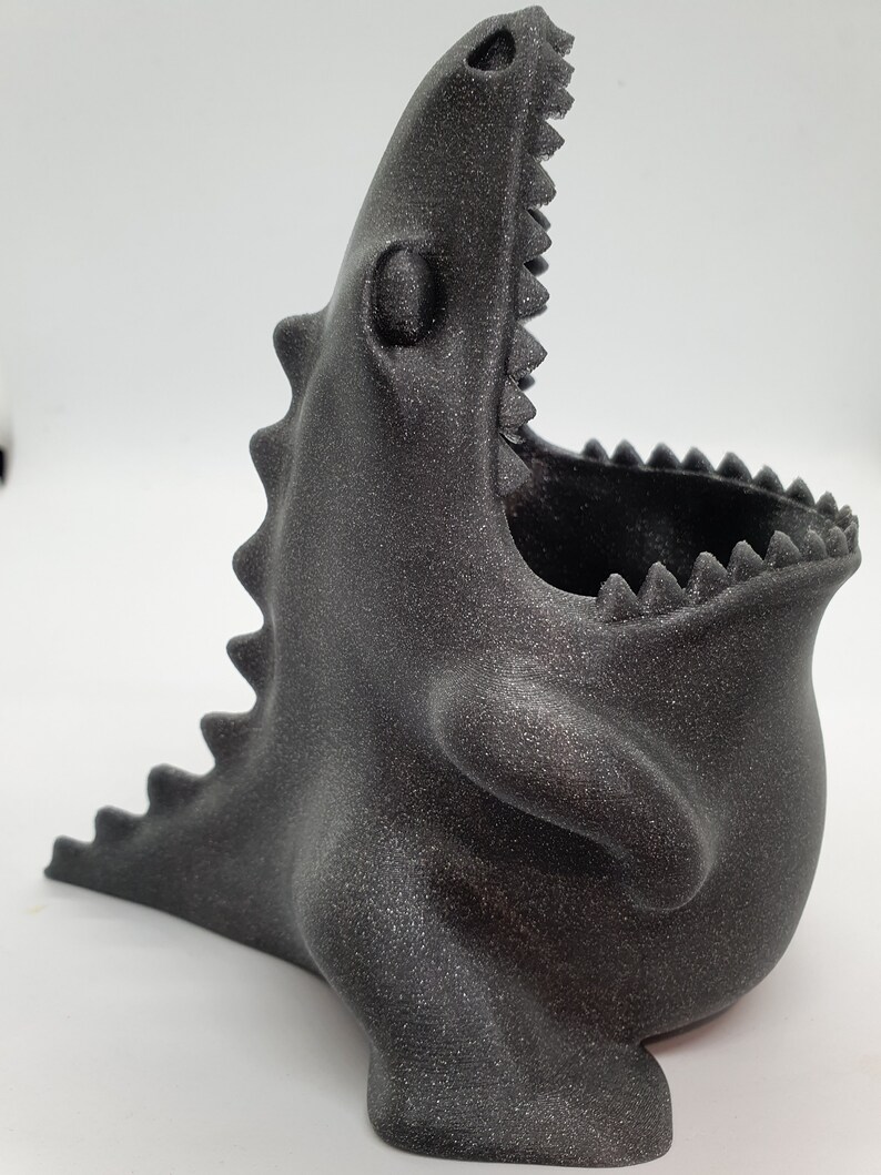 May include: A black, 3D printed dinosaur-shaped planter with a textured surface and an open mouth. The planter is designed to hold small plants or other decorative items.