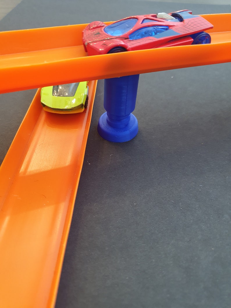 Puede incluir: Dos coches de juguete en un set de pista naranja. La pista superior est&aacute; elevada y sostenida por un pilar de pl&aacute;stico azul. El coche de la pista superior es rojo con un parabrisas azul y detalles negros. El coche de la pista inferior es verde con un parabrisas negro.