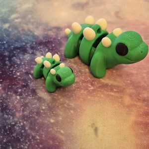 May include: Two 3D printed green dinosaur toys with white spots. The larger dinosaur is segmented and has black eyes. The smaller dinosaur is solid and has black eyes.