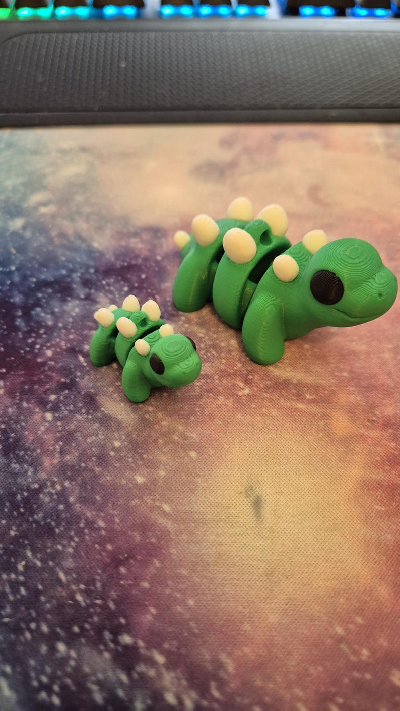 May include: Two 3D printed green dinosaur toys with white spots. The larger dinosaur is segmented and has black eyes. The smaller dinosaur is solid and has black eyes.
