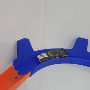 May include: A black toy car with a yellow stripe and silver accents is on a blue plastic track. The track is attached to a white wall. The car is on a curved section of the track.