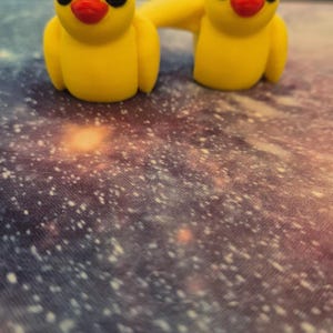 May include: Two yellow rubber duck figurines with black eyes and red beaks. The ducks are connected by a yellow rubber band. The ducks are sitting on a galaxy-patterned surface.