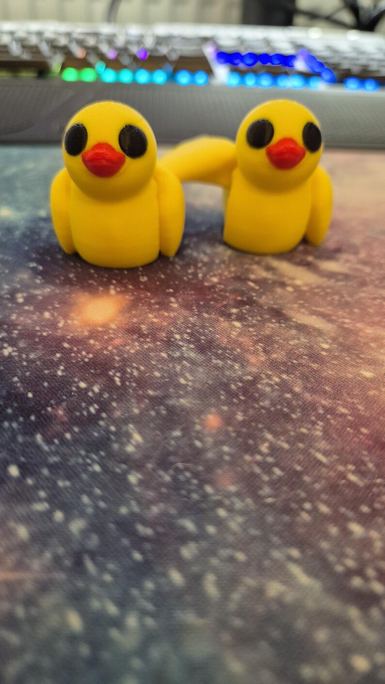 May include: Two yellow rubber duck figurines with black eyes and red beaks. The ducks are connected by a yellow rubber band. The ducks are sitting on a galaxy-patterned surface.