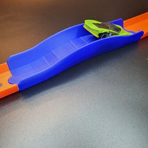 Op de afbeelding: Een blauw plastic baanstuk met een groene speelgoedauto erop. Het baanstuk is gebogen en heeft een getextureerd oppervlak.