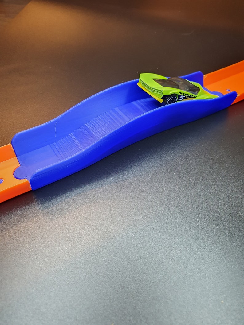 Op de afbeelding: Een blauw plastic baanstuk met een groene speelgoedauto erop. Het baanstuk is gebogen en heeft een getextureerd oppervlak.