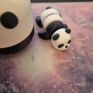 May include: Two panda figurines, one upright and the other lying down. The pandas are black and white, with black ears and eye patches. The lying panda has a segmented body. The background is a galaxy-themed surface.