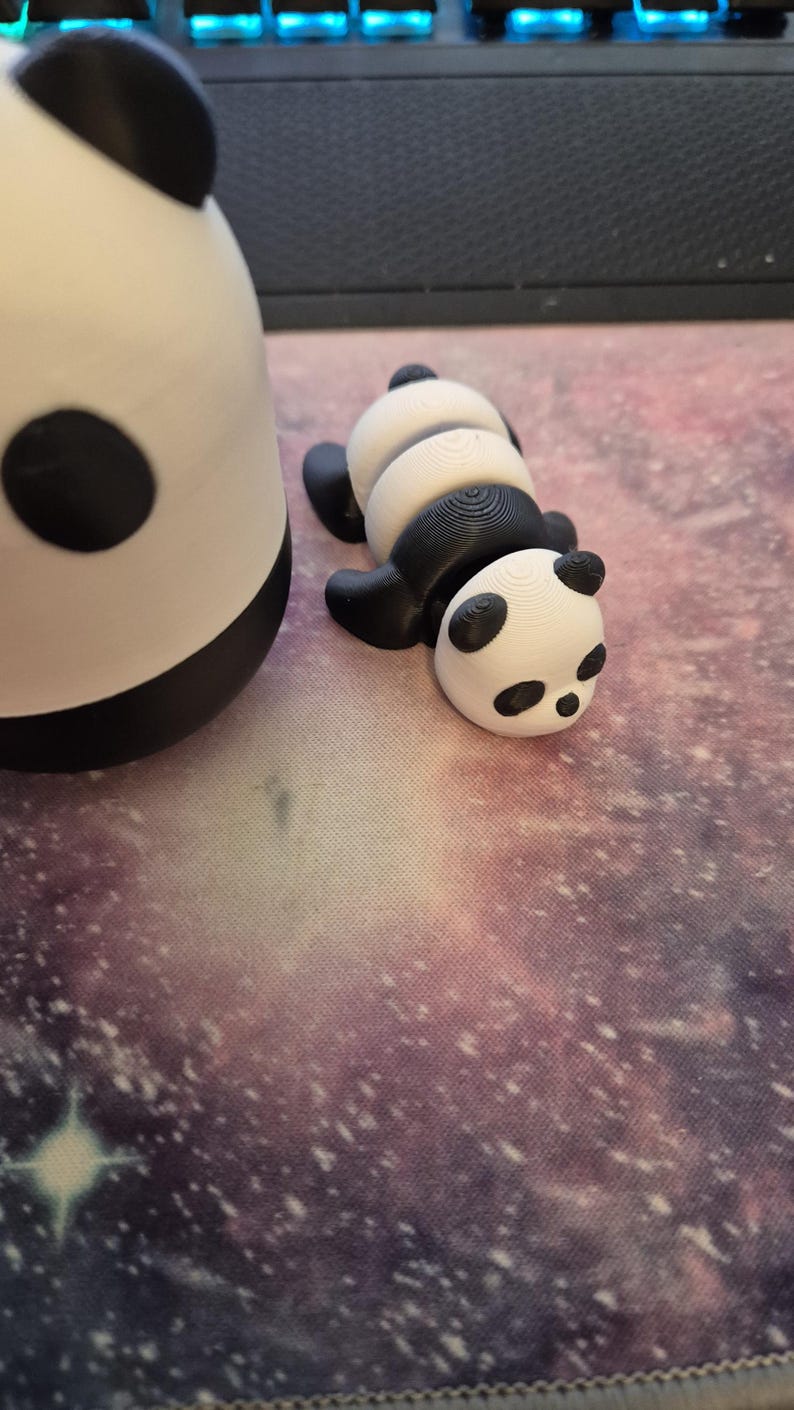 May include: Two panda figurines, one upright and the other lying down. The pandas are black and white, with black ears and eye patches. The lying panda has a segmented body. The background is a galaxy-themed surface.