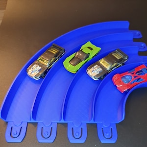 May include: Four toy cars on a blue plastic track. The cars are black, green, red, and silver. The track is curved and has four sections.