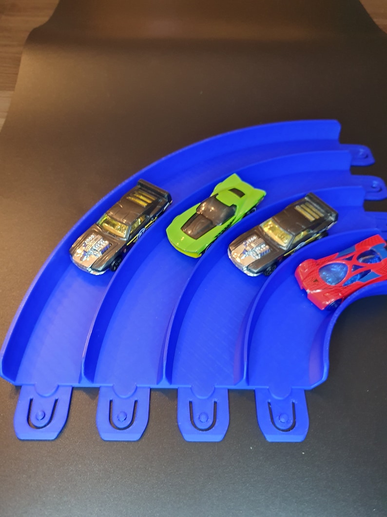 May include: Four toy cars on a blue plastic track. The cars are black, green, red, and silver. The track is curved and has four sections.
