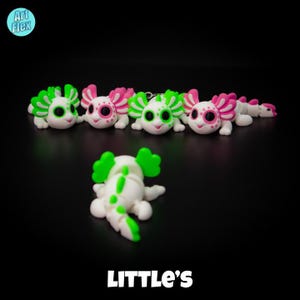 May include: Five white axolotl figurines with green and pink accents. The axolotls are arranged in a row on a black surface. The axolotls have green eyes and are smiling. The text "LITTLE'S" is printed on the surface below the axolotls.