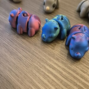 K&ouml;nnte beinhalten: Eine Sammlung kleiner, 3D-gedruckter Tierfiguren in verschiedenen Farben, darunter Grau, Rosa, Blau und Gr&uuml;n. Die Figuren sind auf einer Holzoberfl&auml;che angeordnet.