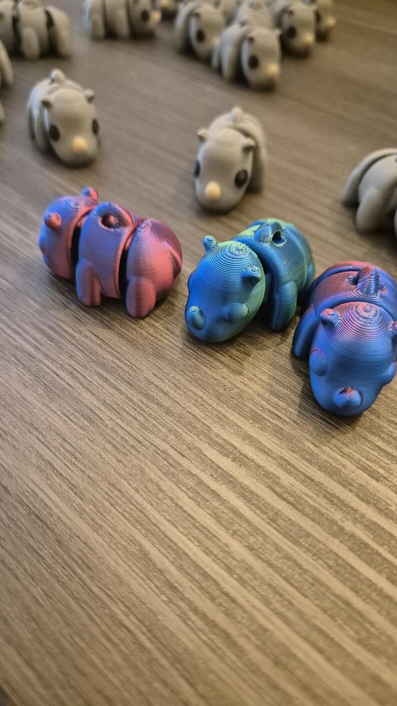 K&ouml;nnte beinhalten: Eine Sammlung kleiner, 3D-gedruckter Tierfiguren in verschiedenen Farben, darunter Grau, Rosa, Blau und Gr&uuml;n. Die Figuren sind auf einer Holzoberfl&auml;che angeordnet.