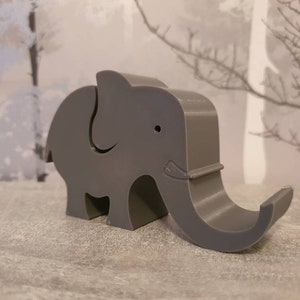 K&ouml;nnte beinhalten: Ein grauer elefantenf&ouml;rmiger Telefonhalter. Der Halter besteht aus einem festen Material und hat einen R&uuml;ssel, der sich nach oben kr&uuml;mmt, um ein Telefon zu halten. Der Elefant hat ein kleines Auge und ein dekoratives Detail an seinem R&uuml;ssel.