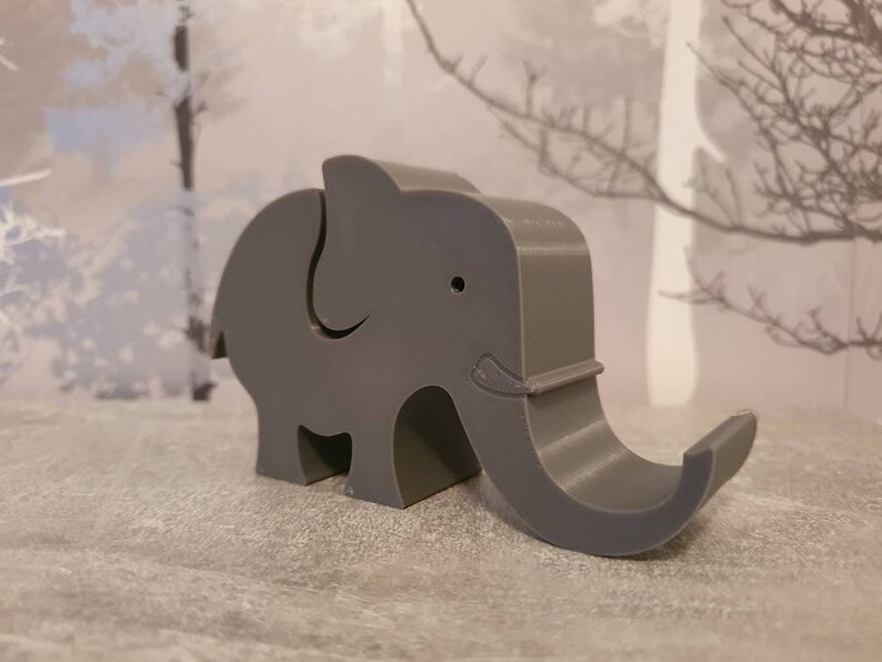 K&ouml;nnte beinhalten: Ein grauer elefantenf&ouml;rmiger Telefonhalter. Der Halter besteht aus einem festen Material und hat einen R&uuml;ssel, der sich nach oben kr&uuml;mmt, um ein Telefon zu halten. Der Elefant hat ein kleines Auge und ein dekoratives Detail an seinem R&uuml;ssel.
