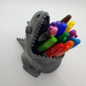 May include: A gray 3D printed dinosaur head pencil holder with a variety of colored pens and markers sticking out of its open mouth.