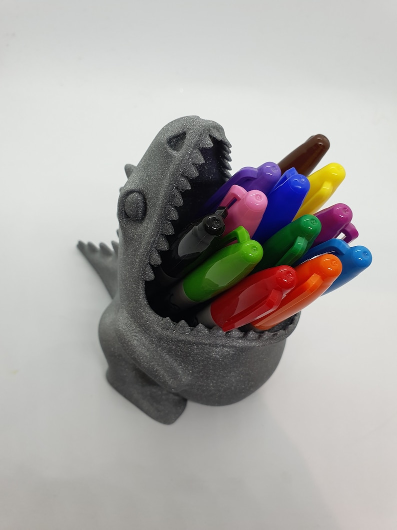 May include: A gray 3D printed dinosaur head pencil holder with a variety of colored pens and markers sticking out of its open mouth.