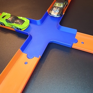 May include: A blue and orange plastic toy race track with two toy cars. One car is green and the other is black.
