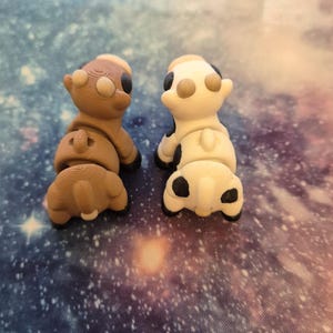 May include: Two small, 3D-printed cow figurines. One is brown with tan and black accents, the other is white with black spots and tan accents. Both are positioned facing away from the viewer, showcasing their rear ends.