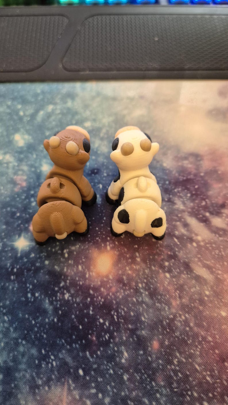 May include: Two small, 3D-printed cow figurines. One is brown with tan and black accents, the other is white with black spots and tan accents. Both are positioned facing away from the viewer, showcasing their rear ends.