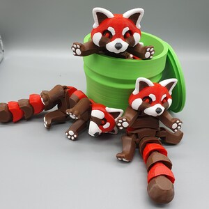 May include: Three red panda figurines, one inside a green container, and two on a gray surface. The red pandas have brown bodies, red and white faces, and curled tails.