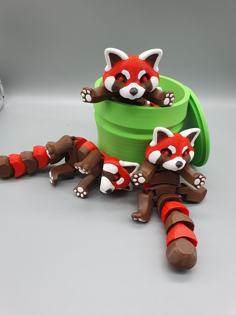 May include: Three red panda figurines, one inside a green container, and two on a gray surface. The red pandas have brown bodies, red and white faces, and curled tails.