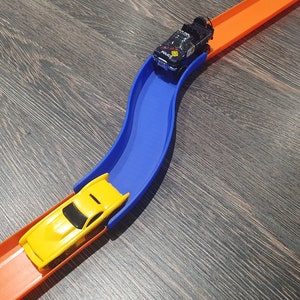 May include: A yellow toy car on an orange track, about to go over a blue plastic ramp. A black toy car is on the orange track above the blue ramp.