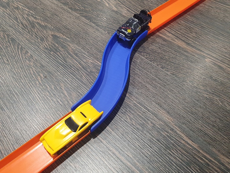May include: A yellow toy car on an orange track, about to go over a blue plastic ramp. A black toy car is on the orange track above the blue ramp.