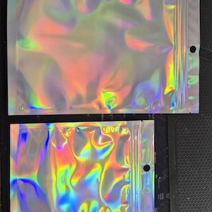 May include: Two iridescent, holographic zip-lock bags. The bags reflect a spectrum of colours, including blues, greens, and yellows. Each bag has a zip closure and a small hole near the top. The bags are made of a shiny, flexible material.
