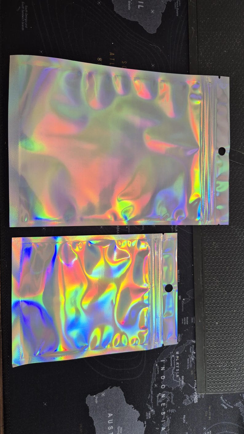 May include: Two iridescent, holographic zip-lock bags. The bags reflect a spectrum of colours, including blues, greens, and yellows. Each bag has a zip closure and a small hole near the top. The bags are made of a shiny, flexible material.