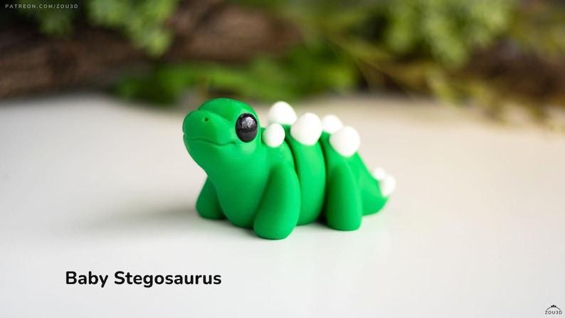 May include: A handcrafted green baby Stegosaurus figurine with white dorsal plates. The dinosaur has large black eyes and is posed on a white surface. The text "Baby Stegosaurus" is at the bottom of the image.