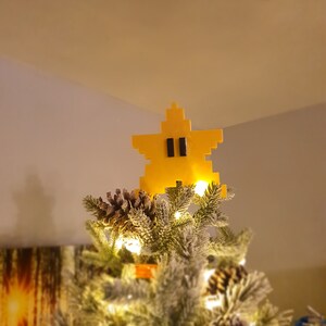 Puede incluir: Un adorno de &aacute;rbol de Navidad en forma de estrella amarilla con ojos negros, que se asemeja a un personaje de videojuego.
