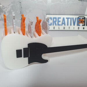 May include: A white guitar-shaped holder with a black pickguard and a black neck. The holder has slots for small figurines, and there are several orange and white figurines in the slots.