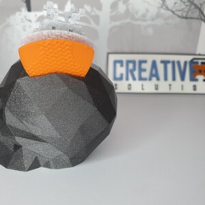 Puede incluir: Un cr&aacute;neo negro impreso en 3D con un dise&ntilde;o geom&eacute;trico. Una peque&ntilde;a nave espacial impresa en 3D de color naranja y blanco se encuentra en la parte superior del cr&aacute;neo. El fondo es una imagen borrosa de una rama de &aacute;rbol y un letrero que dice "CREATIVE 3D SOLUTIONS".